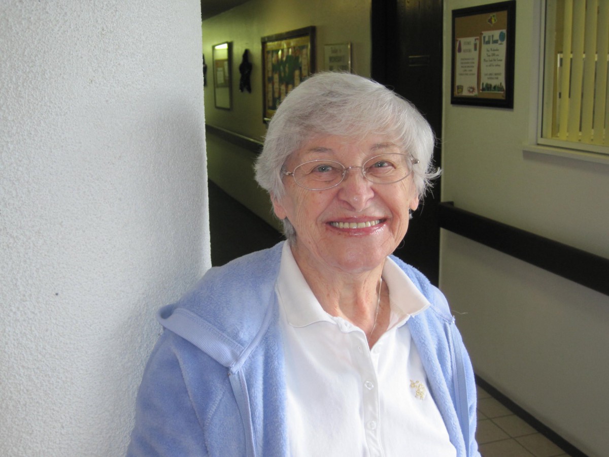An elderly woman smiling while standing indoors, dressed in casual clothing.