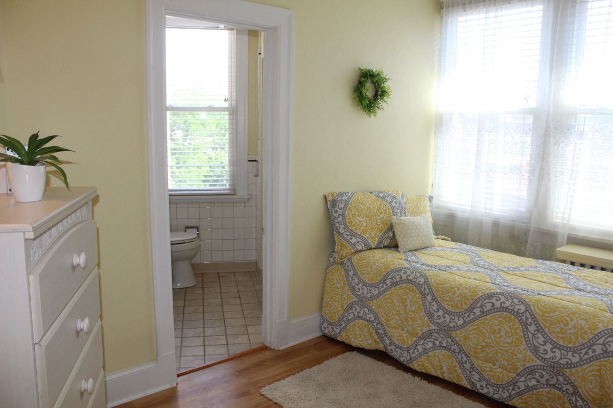 A cozy bedroom with a yellow-patterned daybed next to a small bathroom, illuminated by natural light from the window.