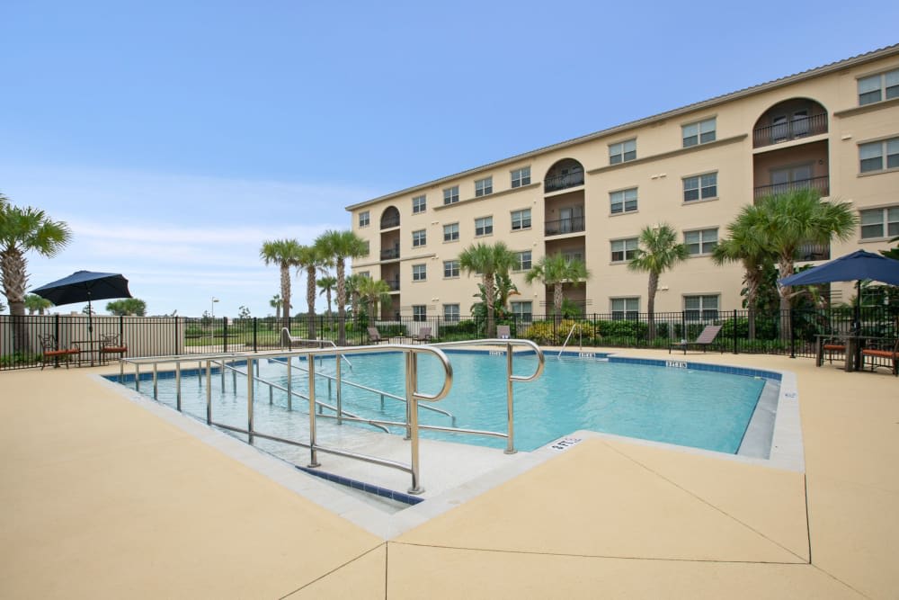 An outdoor swimming pool in front of an apartment building, surrounded by palm trees under a clear sky.