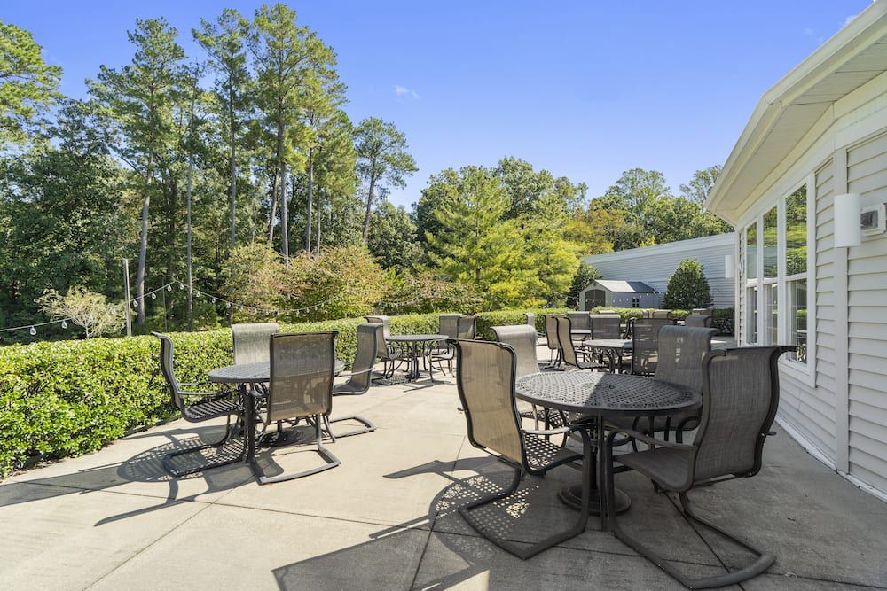 A spacious outdoor patio area with multiple tables and chairs surrounded by lush greenery and trees under a clear blue sky.