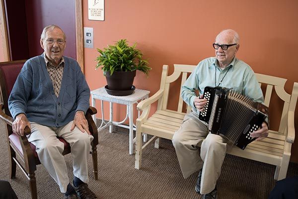 Two elderly men, one playing an accordion and the other seated beside him, enjoy a moment indoors next to a potted plant.