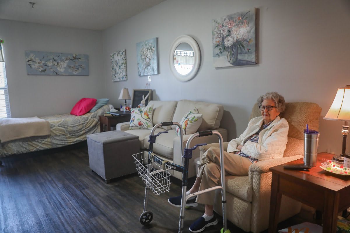 An elderly woman sitting in a cozy living room with a sofa and walker nearby.