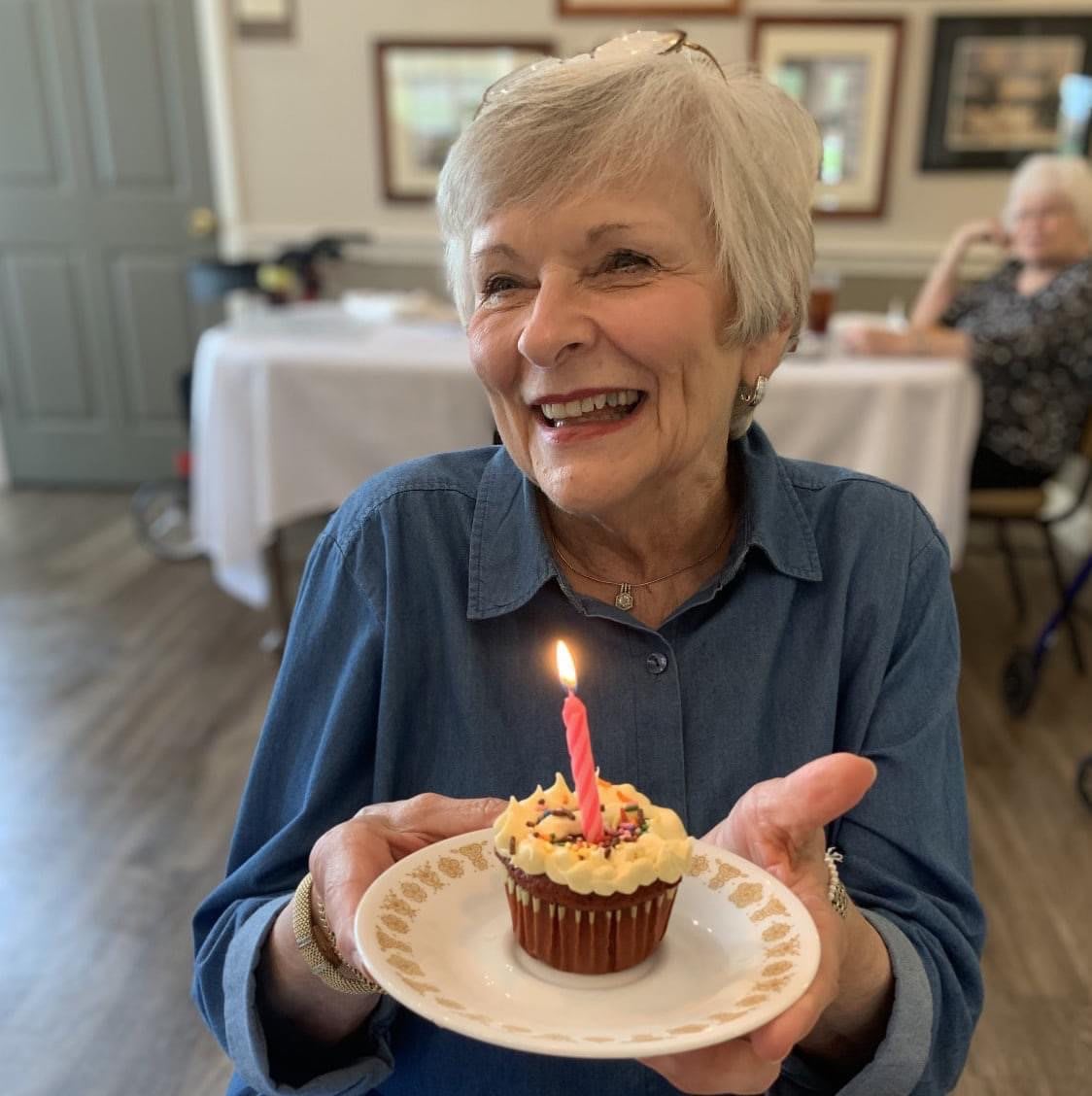 An elderly woman smiling and holding a cupcake with a lit candle, celebrating her birthday.