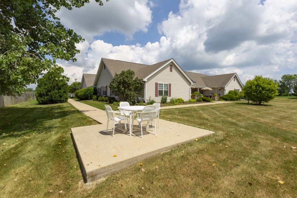 A suburban house with a small patio area featuring outdoor furniture on a grassy lawn under a cloudy sky.