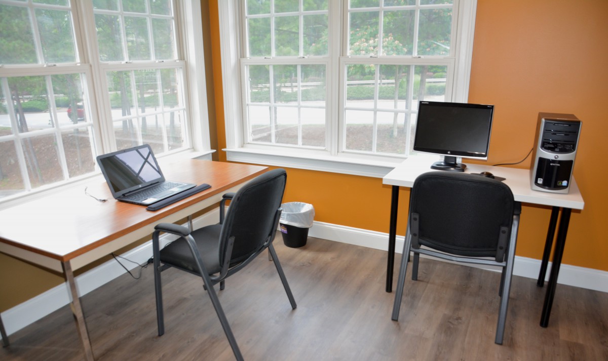 A bright office workspace featuring desks, computers, and chairs near large windows.