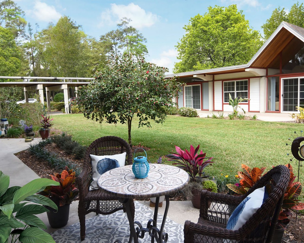 A serene garden patio featuring wicker chairs and a round table set against a backdrop of lush greenery.