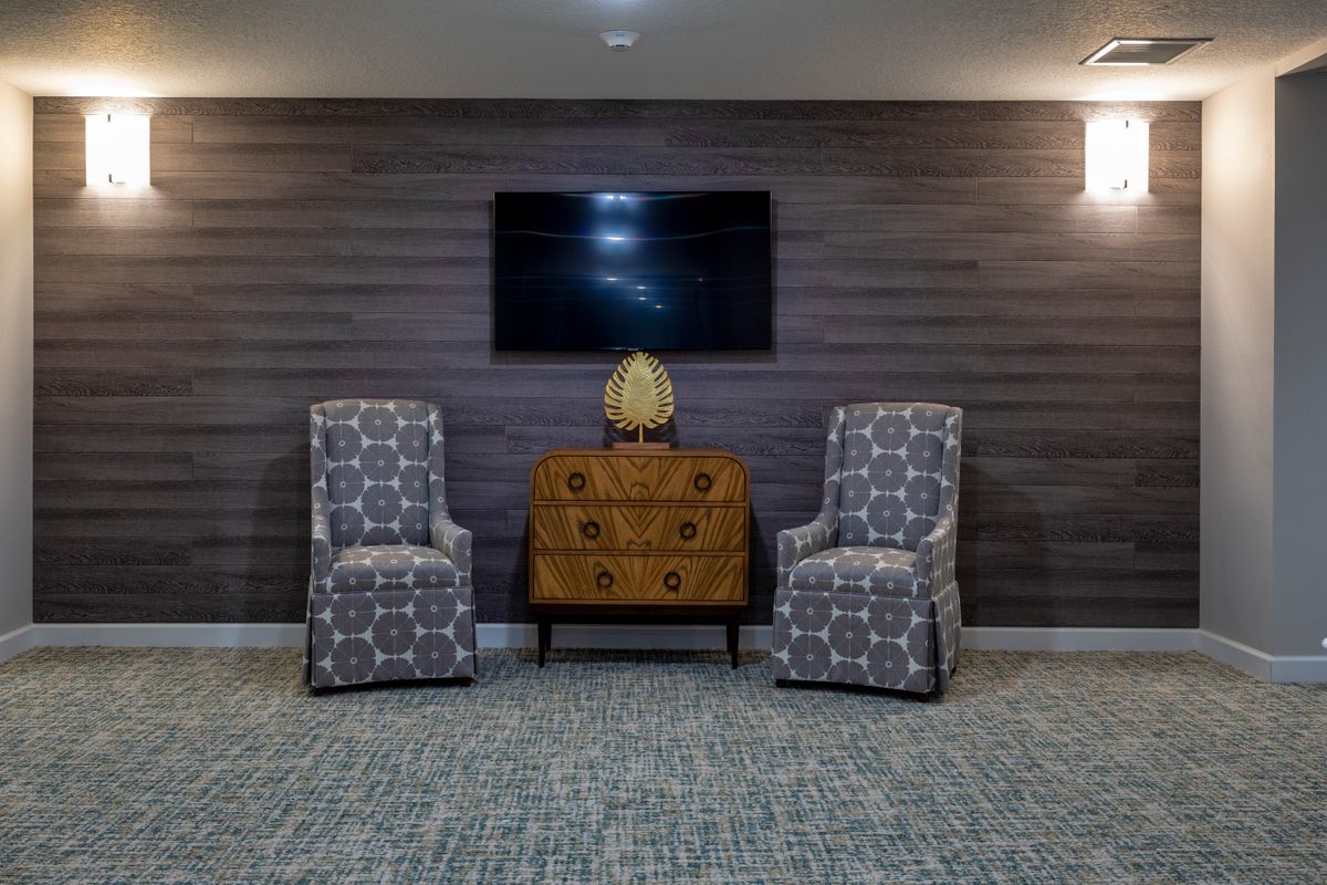 A cozy living room setup featuring two patterned chairs flanking a wooden cabinet with a decorative piece, all set against a dark paneled wall with a mounted television.