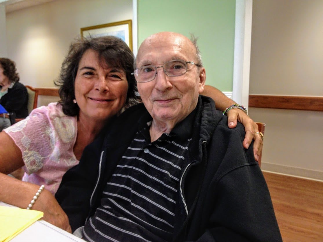 A woman and an elderly man sitting together and smiling indoors.