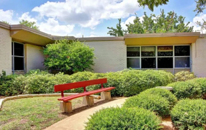A tranquil outdoor scene featuring a red bench surrounded by greenery and a modern building in the background.