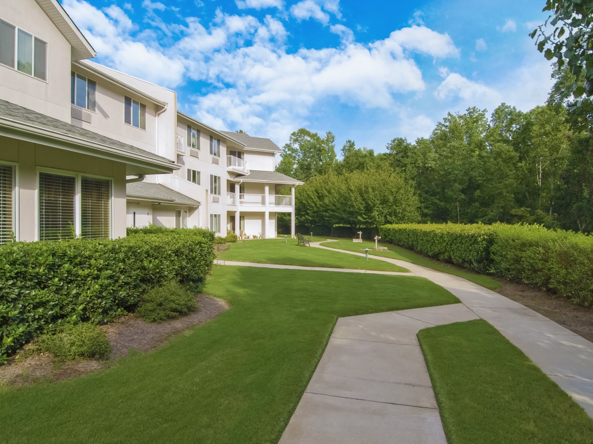 A residential building with a well-maintained lawn and sidewalk, surrounded by trees under a partly cloudy sky.
