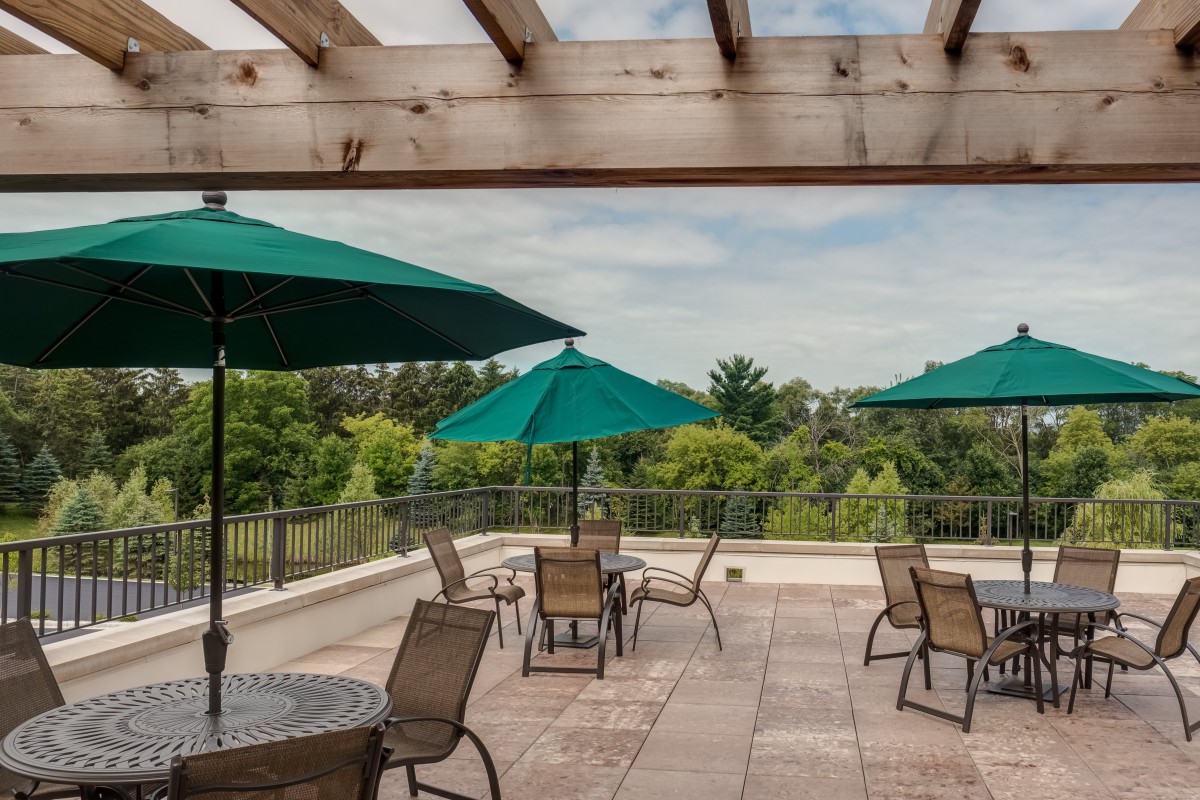 A rooftop patio with tables, chairs, and green umbrellas under a pergola overlooking lush greenery.