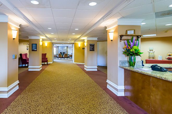 A brightly lit hotel lobby with a carpeted hallway, reception area, and decorative flowers.