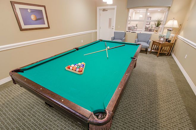 A billiards table with a green felt top and a triangular rack of pool balls, located in a recreation room with chairs in the background.