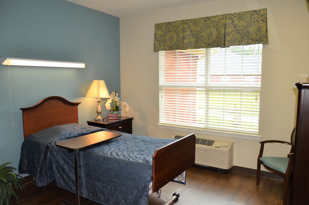 A hospital room with a single bed, nightstand, lamp, and window.
