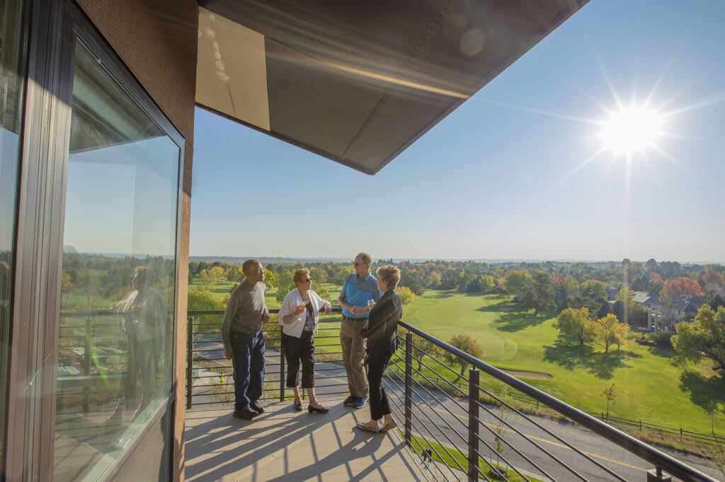 A group of people stand on a balcony overlooking a golf course on a sunny day.