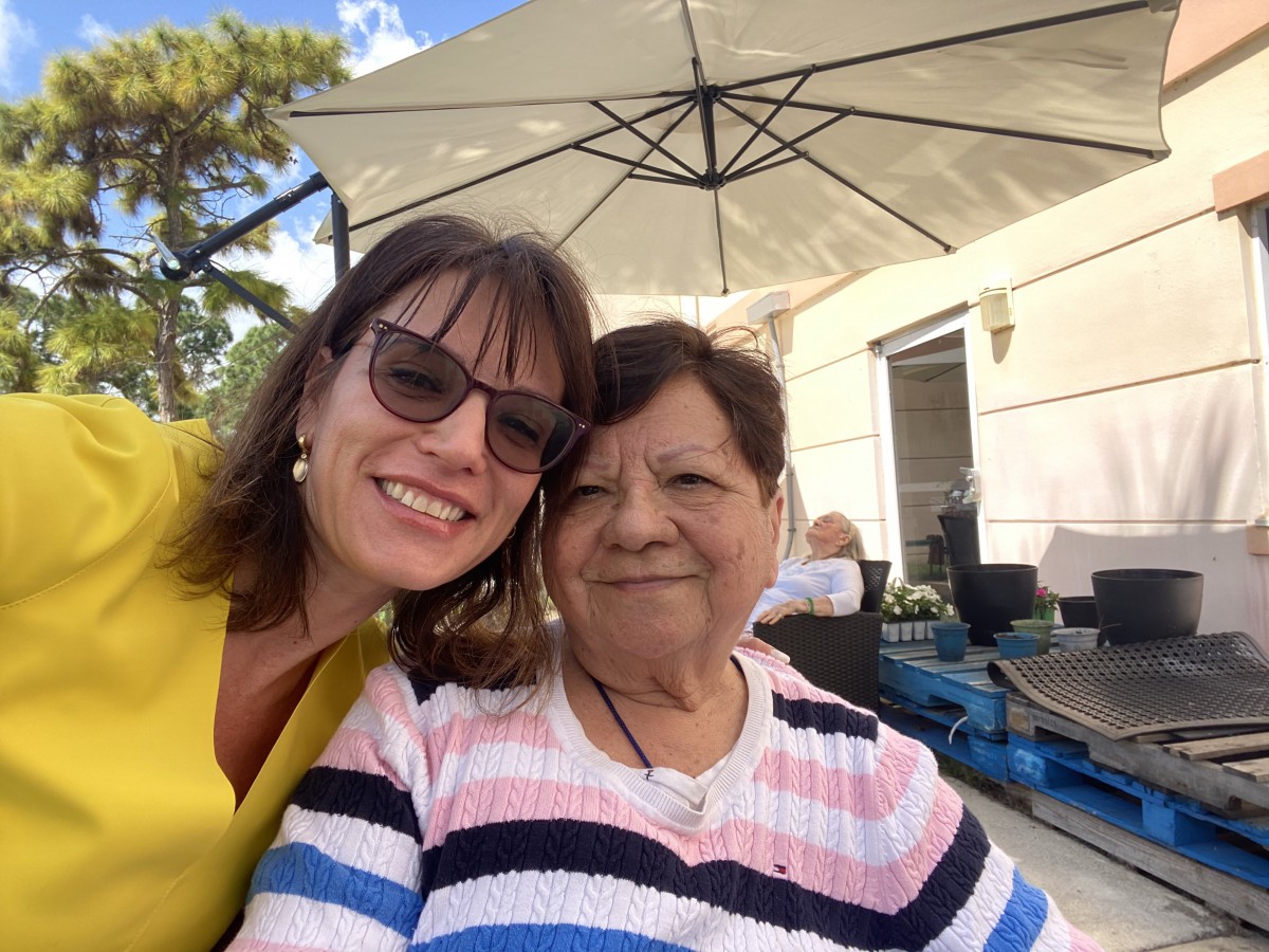 Two people smiling while taking a selfie outdoors under a large umbrella.