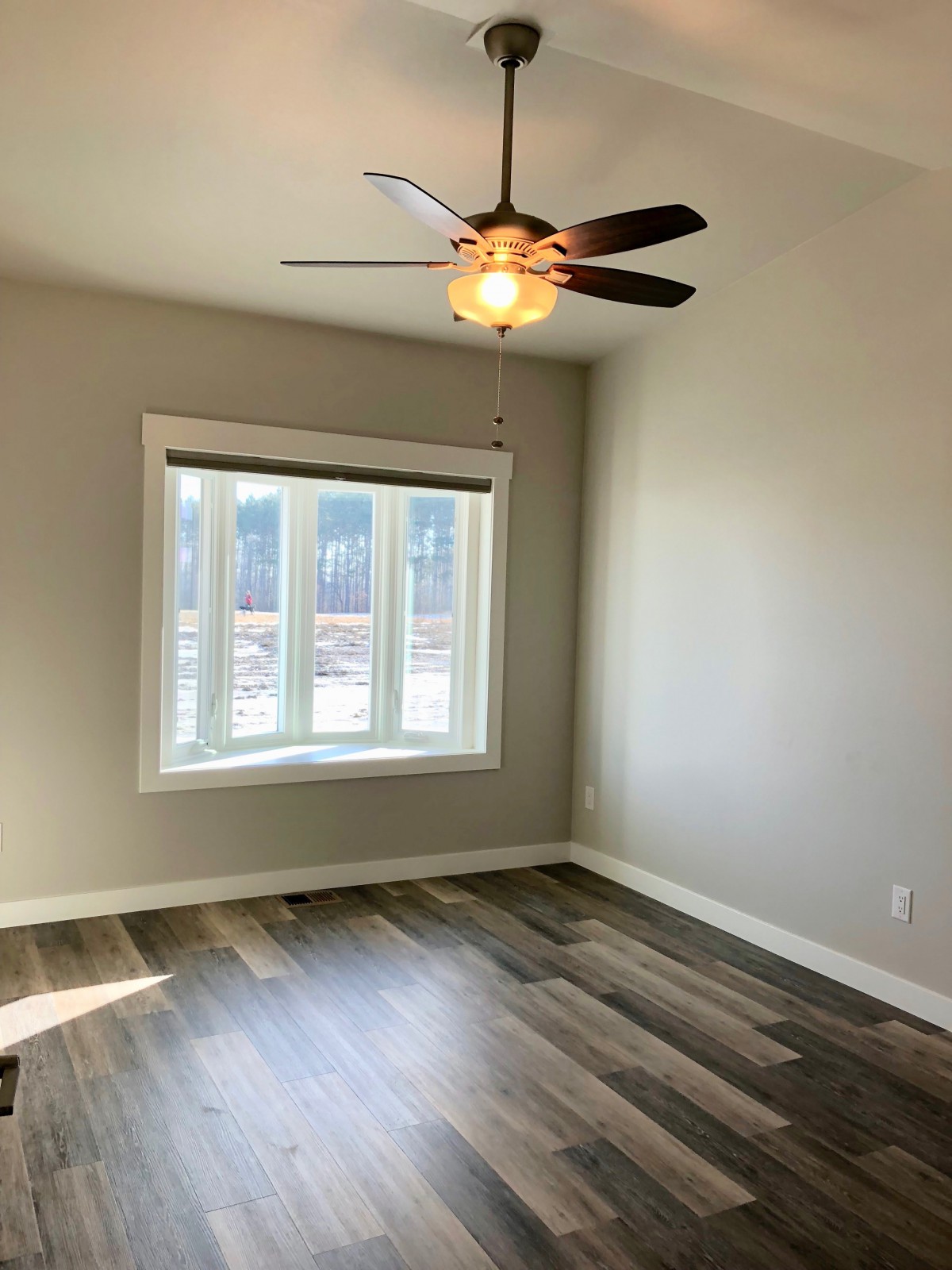 An empty room with a ceiling fan and large window, featuring hardwood floors.