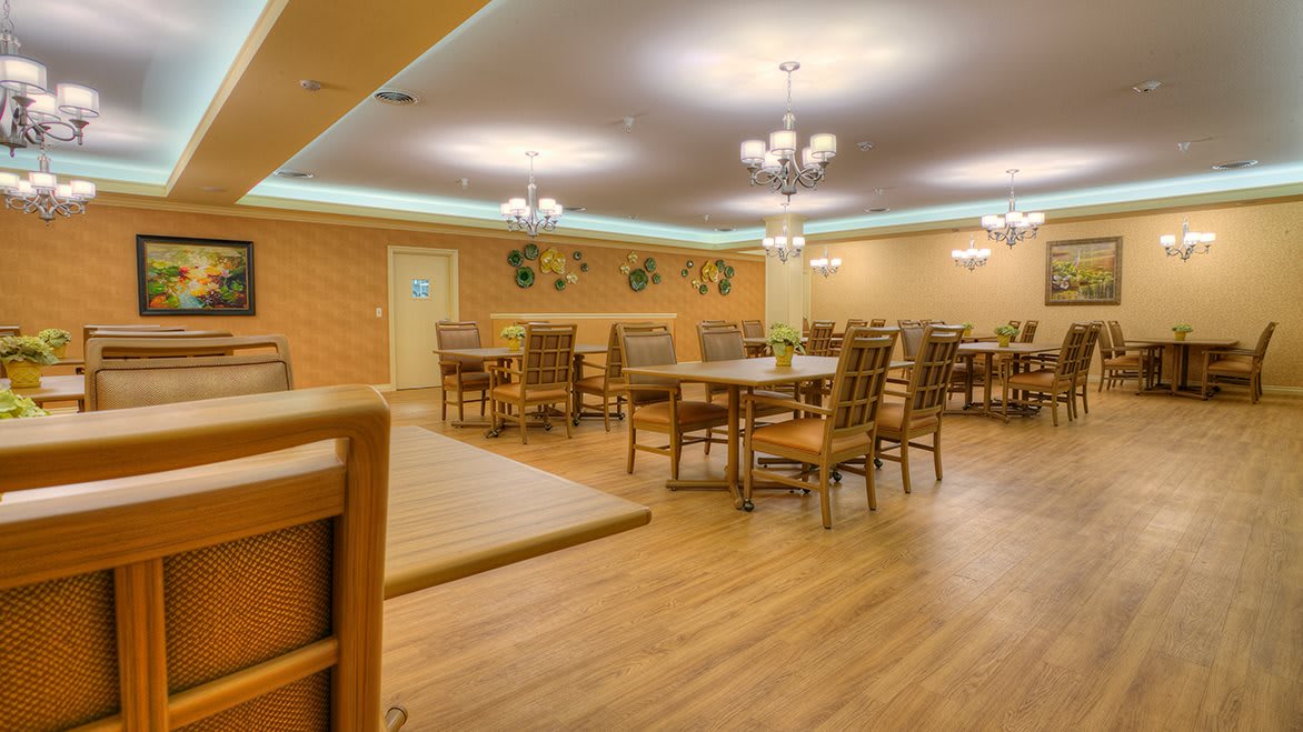 A spacious dining room with wooden floors, featuring several tables and chairs under elegant chandeliers.