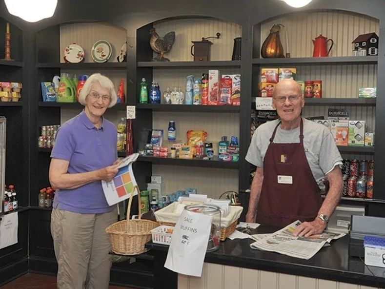 Two people in a small grocery store, one holding a basket and the other standing behind the counter.
