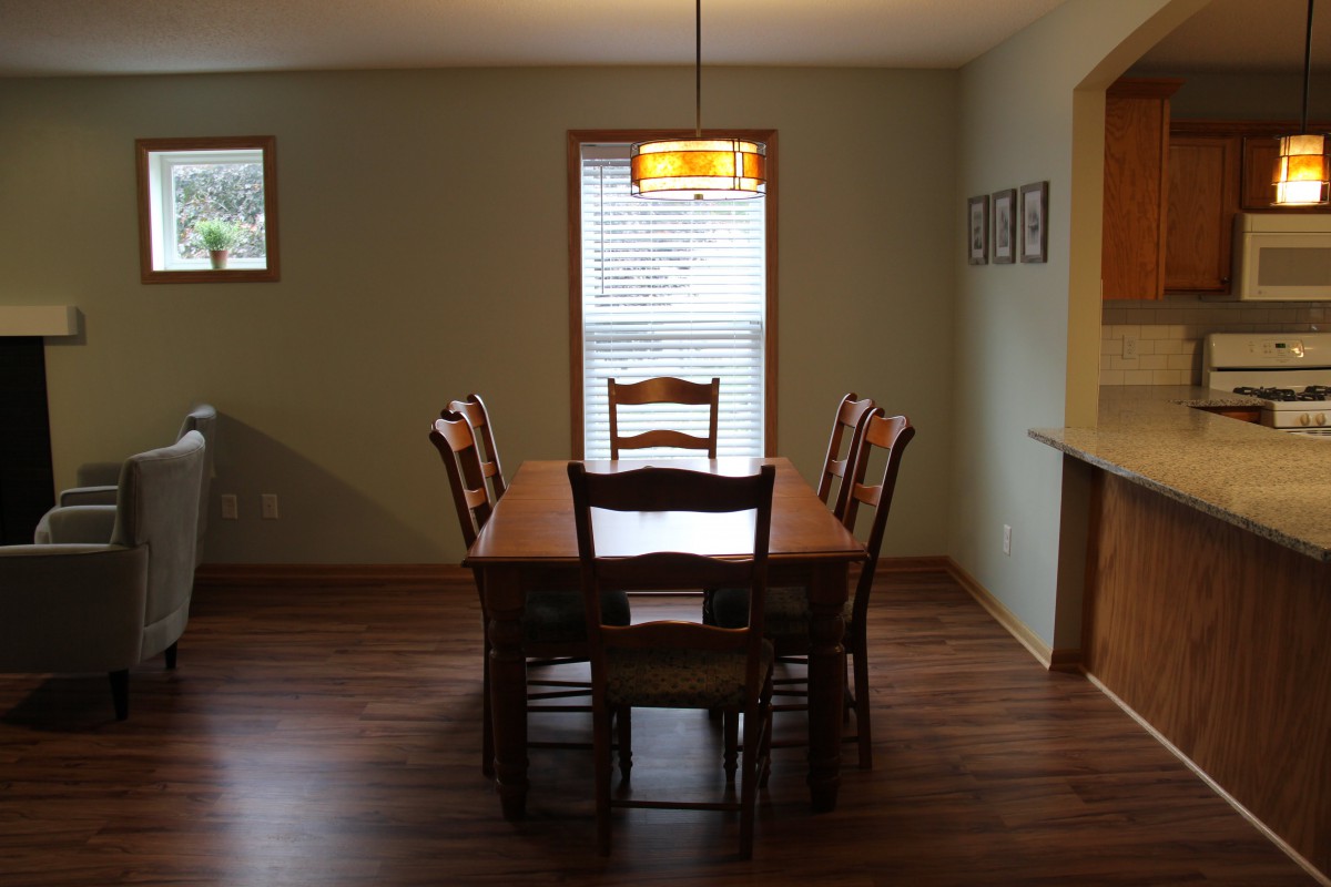 A cozy dining room with a wooden table and chairs, featuring a central light fixture and large window.