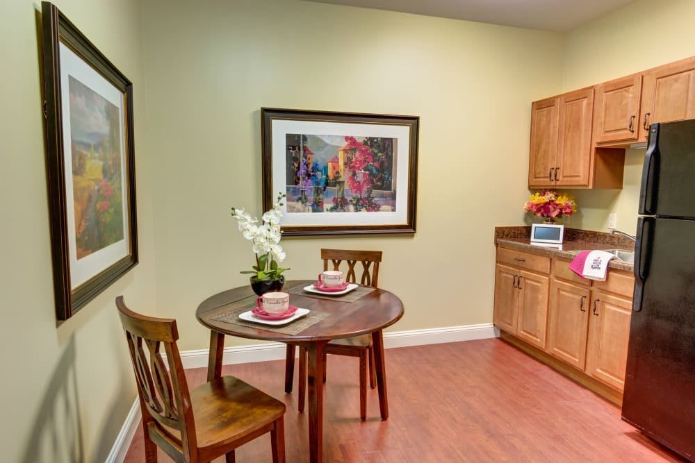 A cozy kitchenette with a round table set for two, featuring wooden chairs, a decorative orchid centerpiece, and framed artwork on a light green wall.