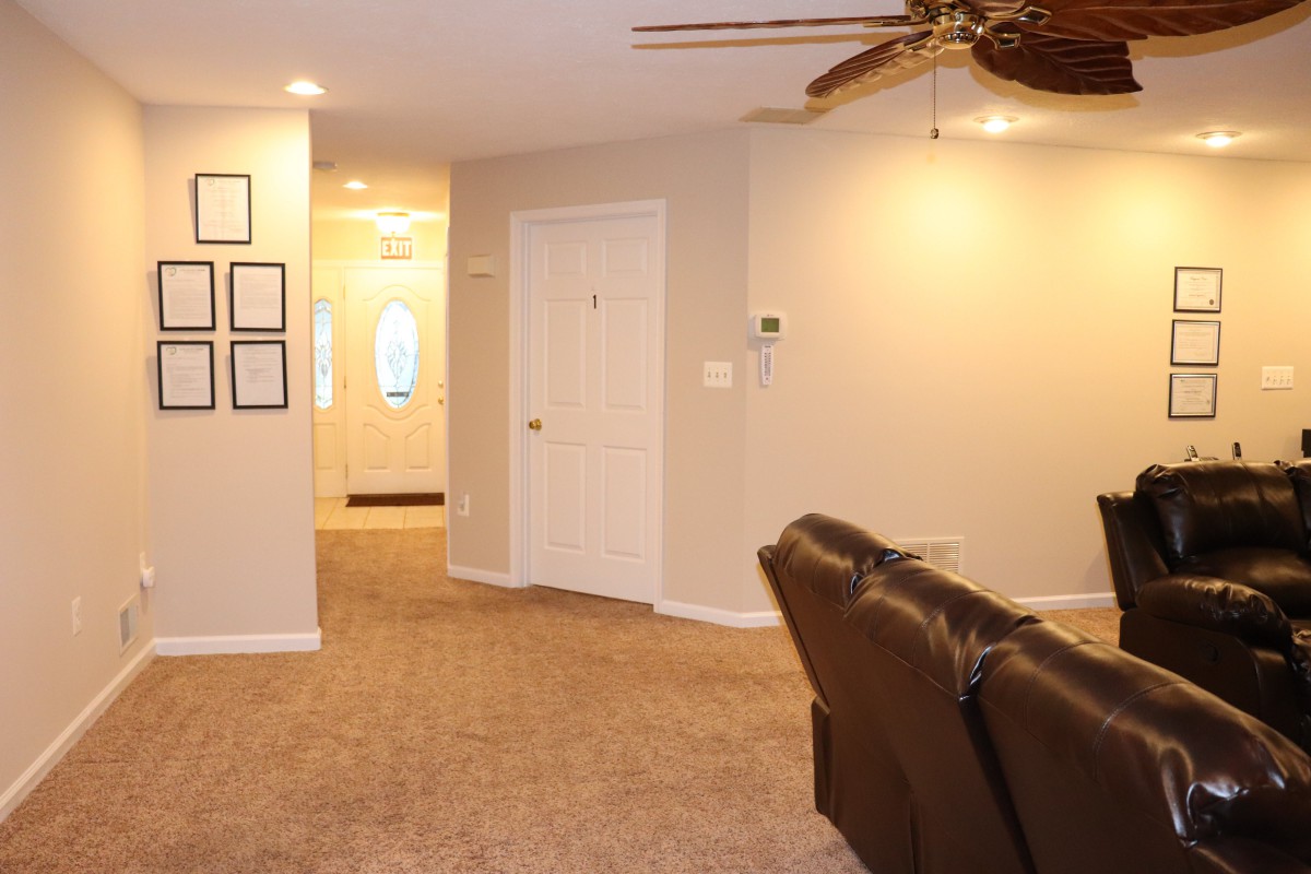 A cozy living room with carpet flooring, a ceiling fan, and a comfortable recliner leading to a bright hallway.