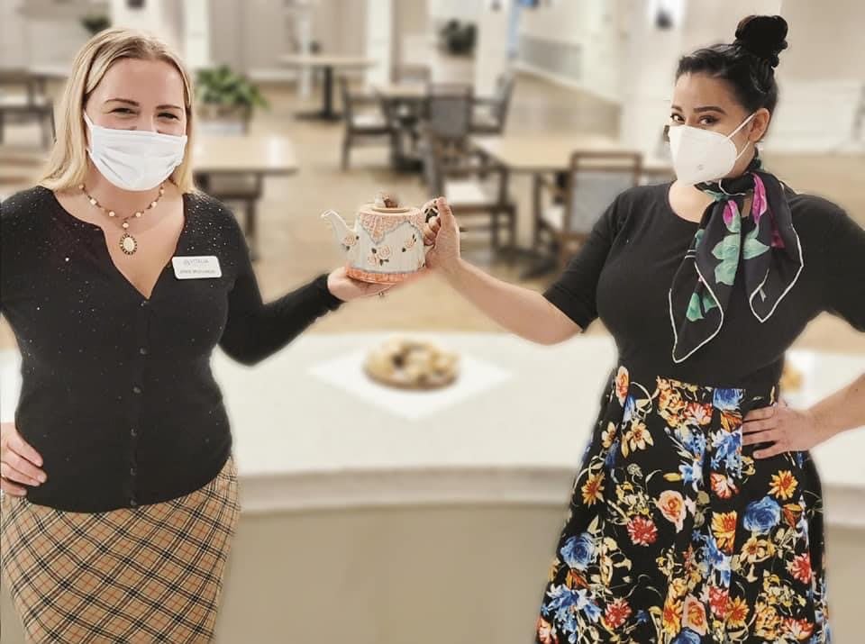 Two women indoors wearing masks, holding a teapot together in a social setting.