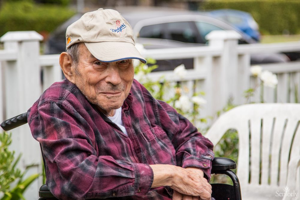 An elderly man wearing a plaid shirt and cap sits outdoors in a wheelchair.