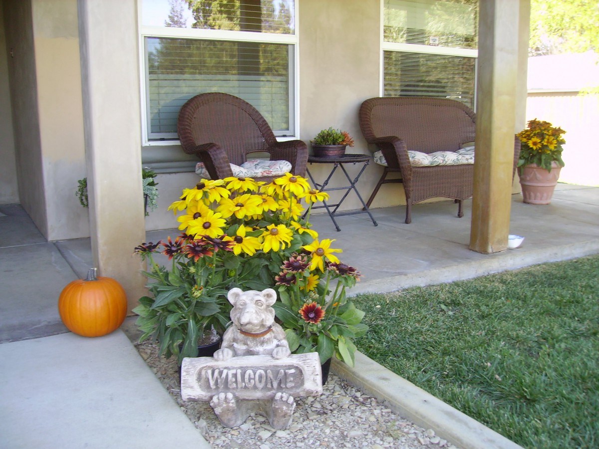 A cozy front porch with wicker chairs, vibrant yellow flowers, a pumpkin, and a welcome sign statue.