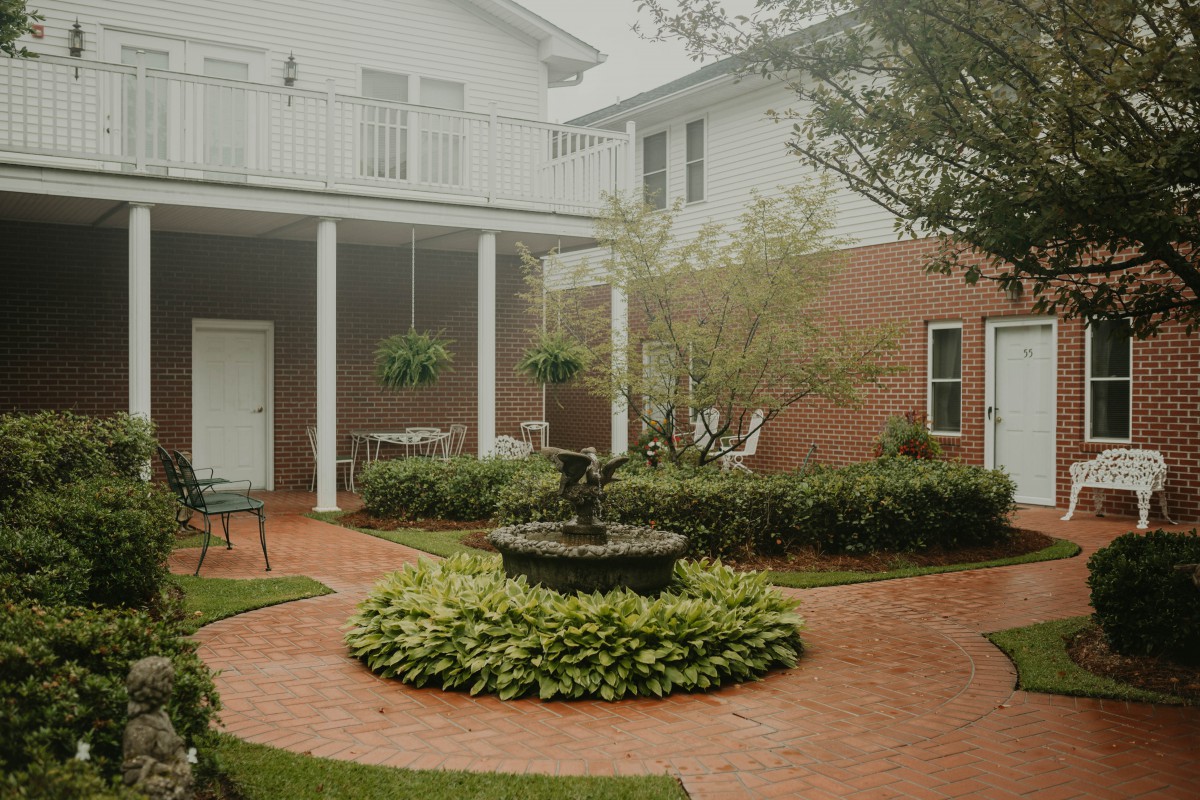 A serene courtyard featuring a central fountain, surrounded by lush greenery and a brick pathway.