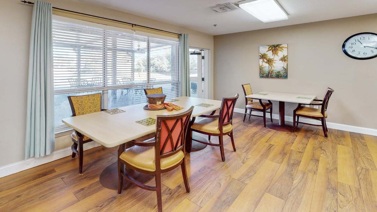 A well-lit dining room with wooden floors, featuring several tables and chairs, and bright natural light from large windows.
