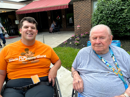 Two individuals enjoying a sunny day outdoors, both smiling and seated in wheelchairs.