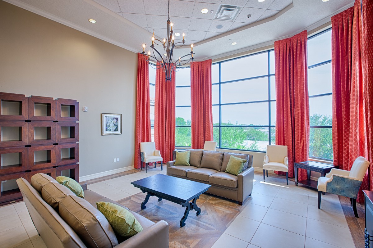 A modern living room with spacious seating, featuring a chandelier and large windows adorned with red curtains.