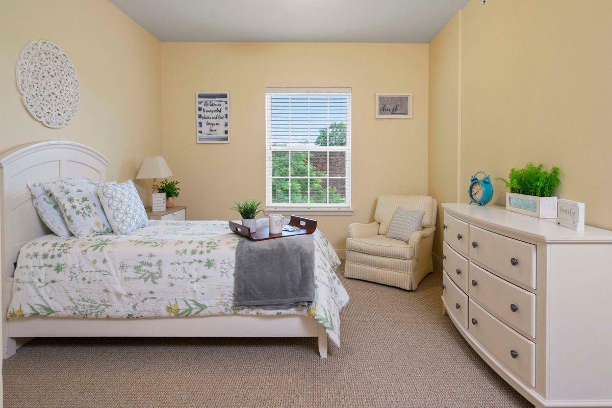 A cozy bedroom with a bed, chair, and dresser, featuring soft lighting and decor.