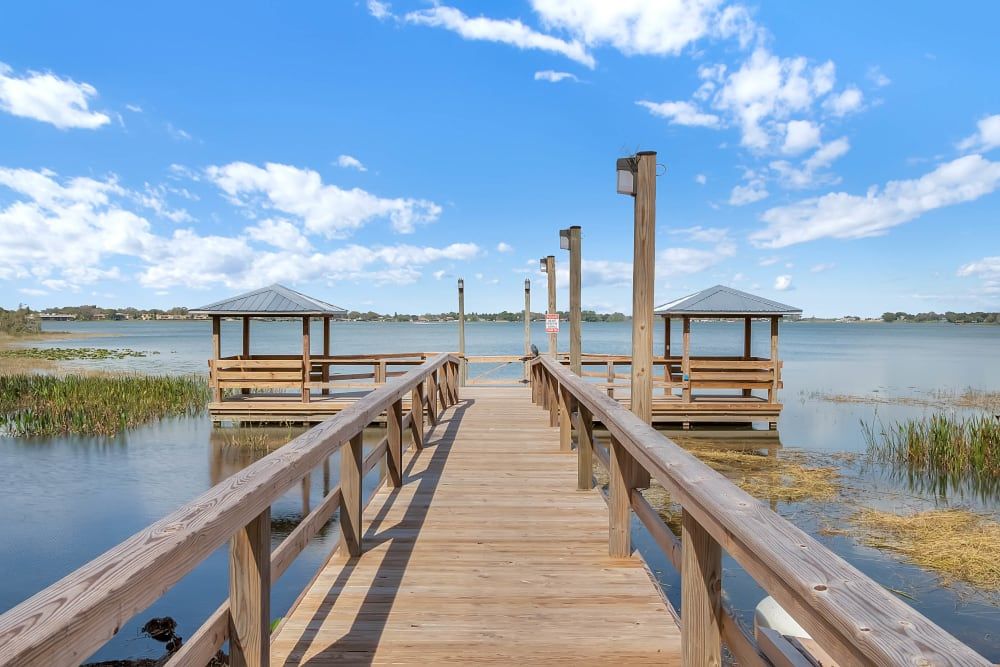 A scenic view of a wooden pier extending into a serene lake, flanked by gazebos under a bright blue sky with scattered clouds.
