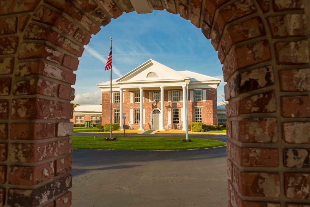 A colonial-style building framed by a brick archway, featuring white pillars and an American flag in the foreground.