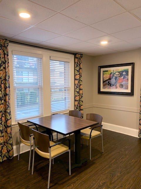 A modern dining area with a wooden table, four chairs, large windows with patterned curtains, and framed artwork on the wall.