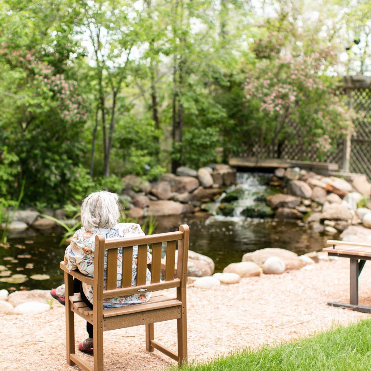 A person sits on a bench by a pond with a small waterfall in a tranquil park setting.