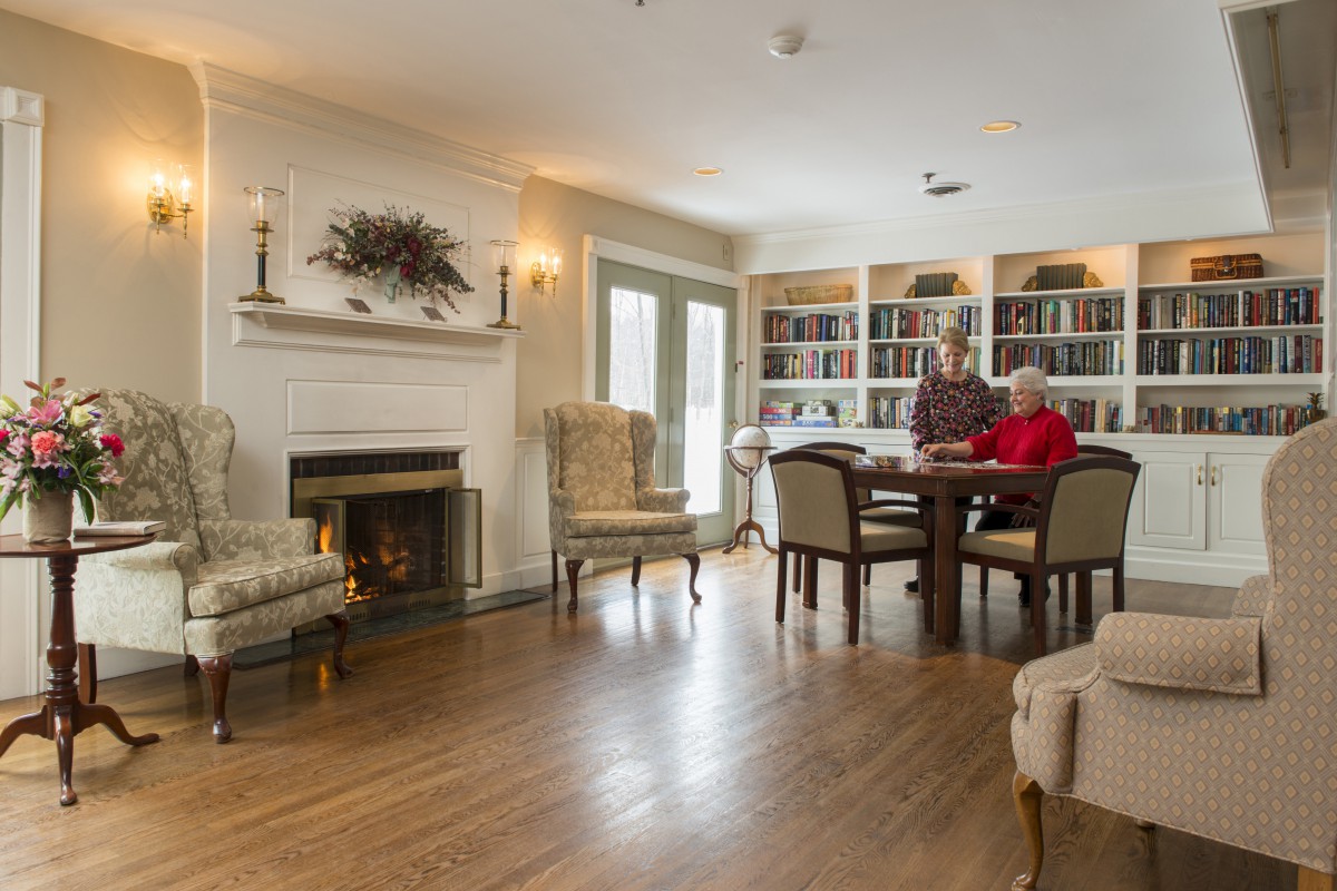 A cozy living room with a fireplace, bookshelves, and two people sitting at a table.
