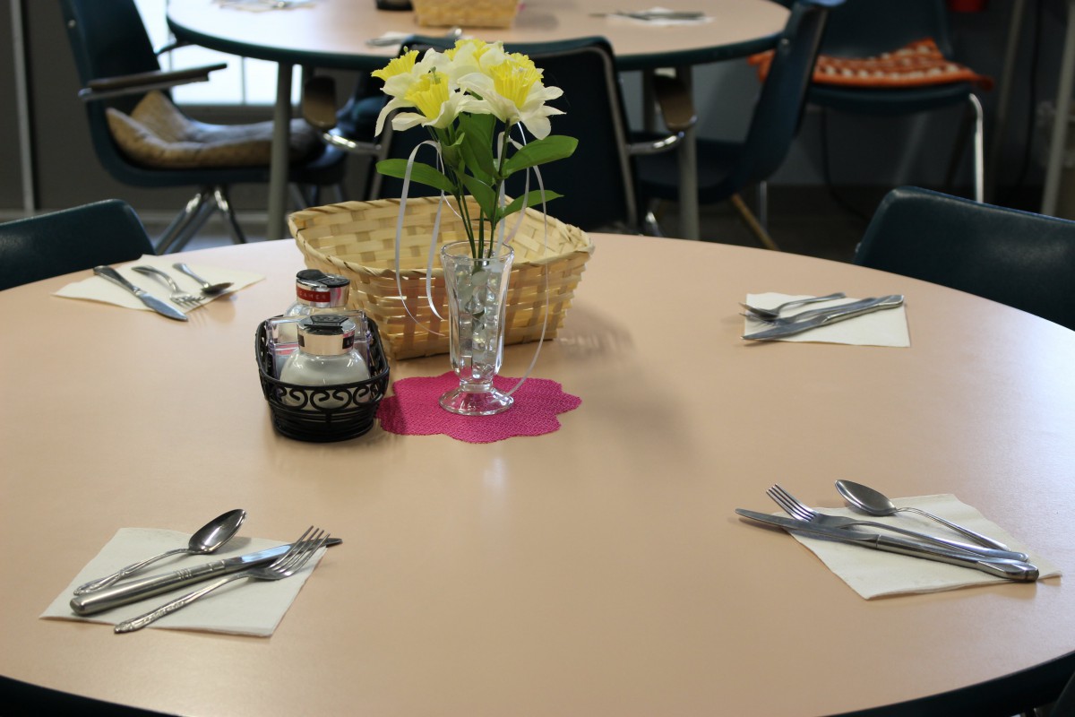 A dining table is set with silverware, a bottle of syrup, and a vase of flowers.
