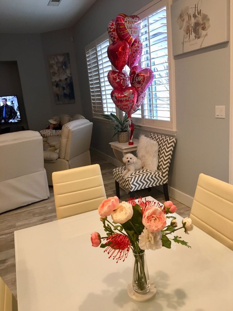 A cozy living and dining area with a floral centerpiece, heart-shaped balloons, and a small dog sitting on a chair.