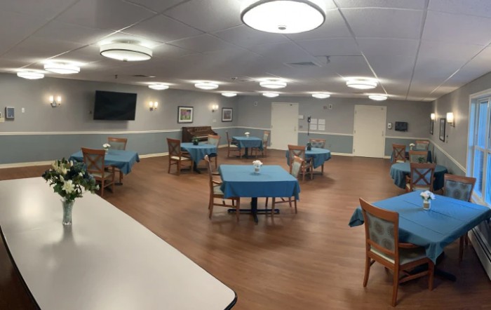 A spacious dining area with neatly arranged tables and chairs, featuring soft lighting and a television on the wall.
