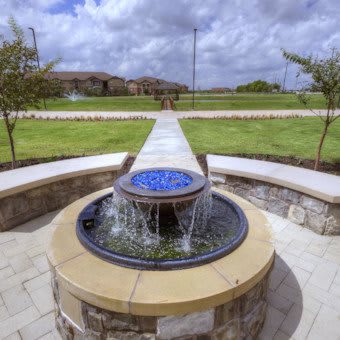 A beautifully designed outdoor fountain with a stone base, set in a landscaped garden with a pathway leading into the distance.