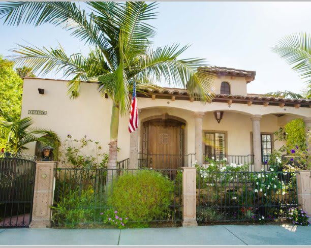A residential house with palm trees and an American flag at the front, surrounded by a garden and a fence.