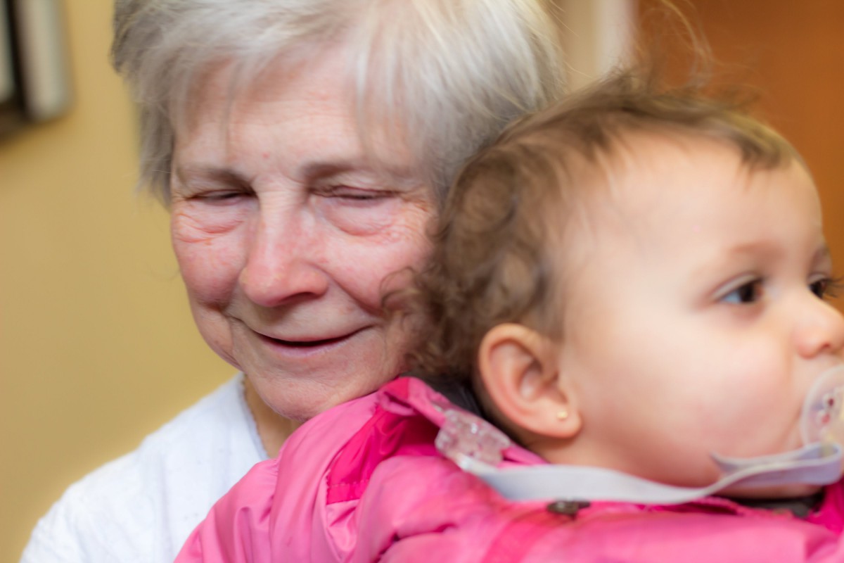 An elderly woman smiling while holding a baby, showcasing a moment of family togetherness.