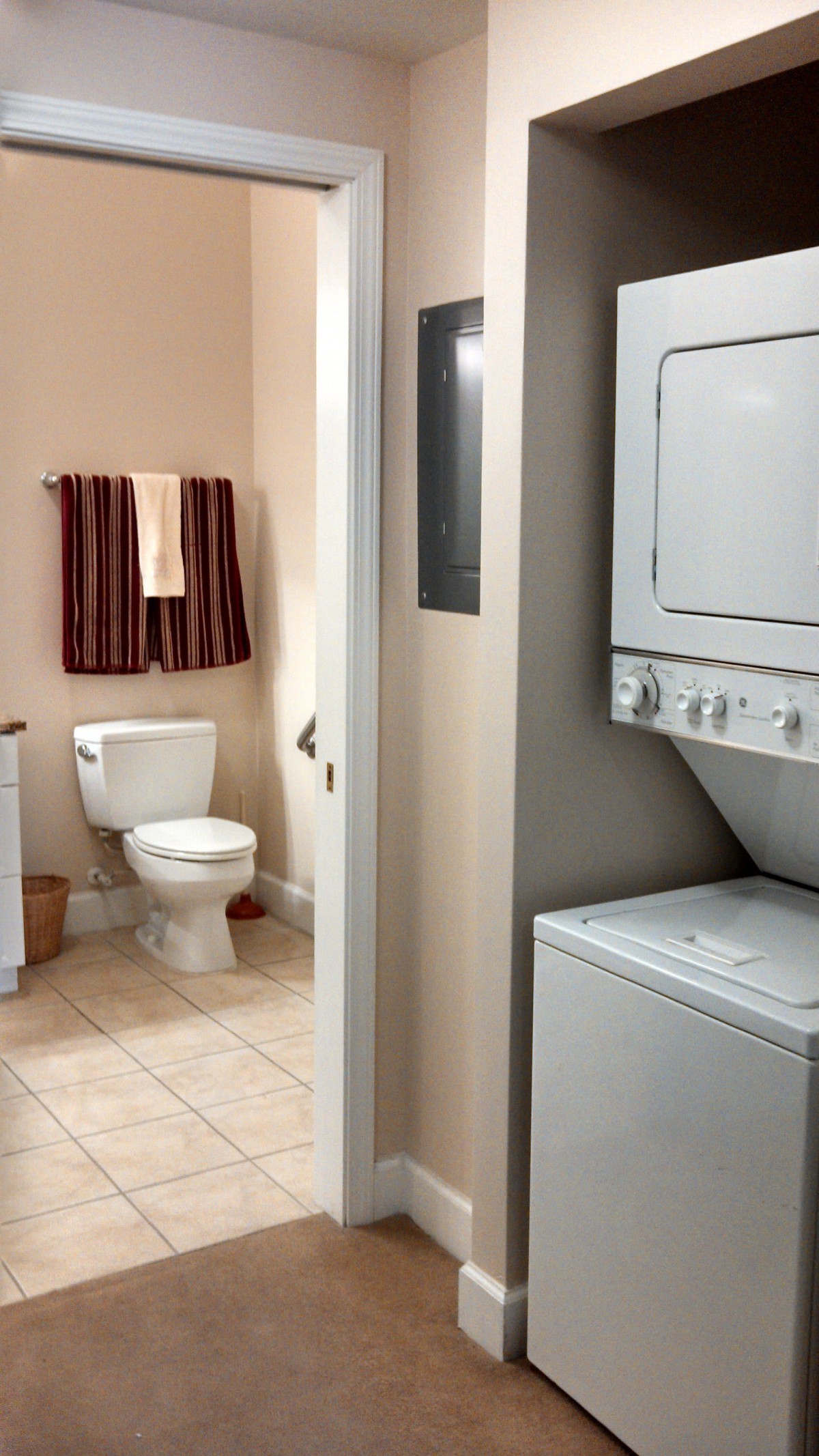A compact dual-purpose room featuring a bathroom with a toilet and towels, alongside a laundry area with a stacked washer and dryer.