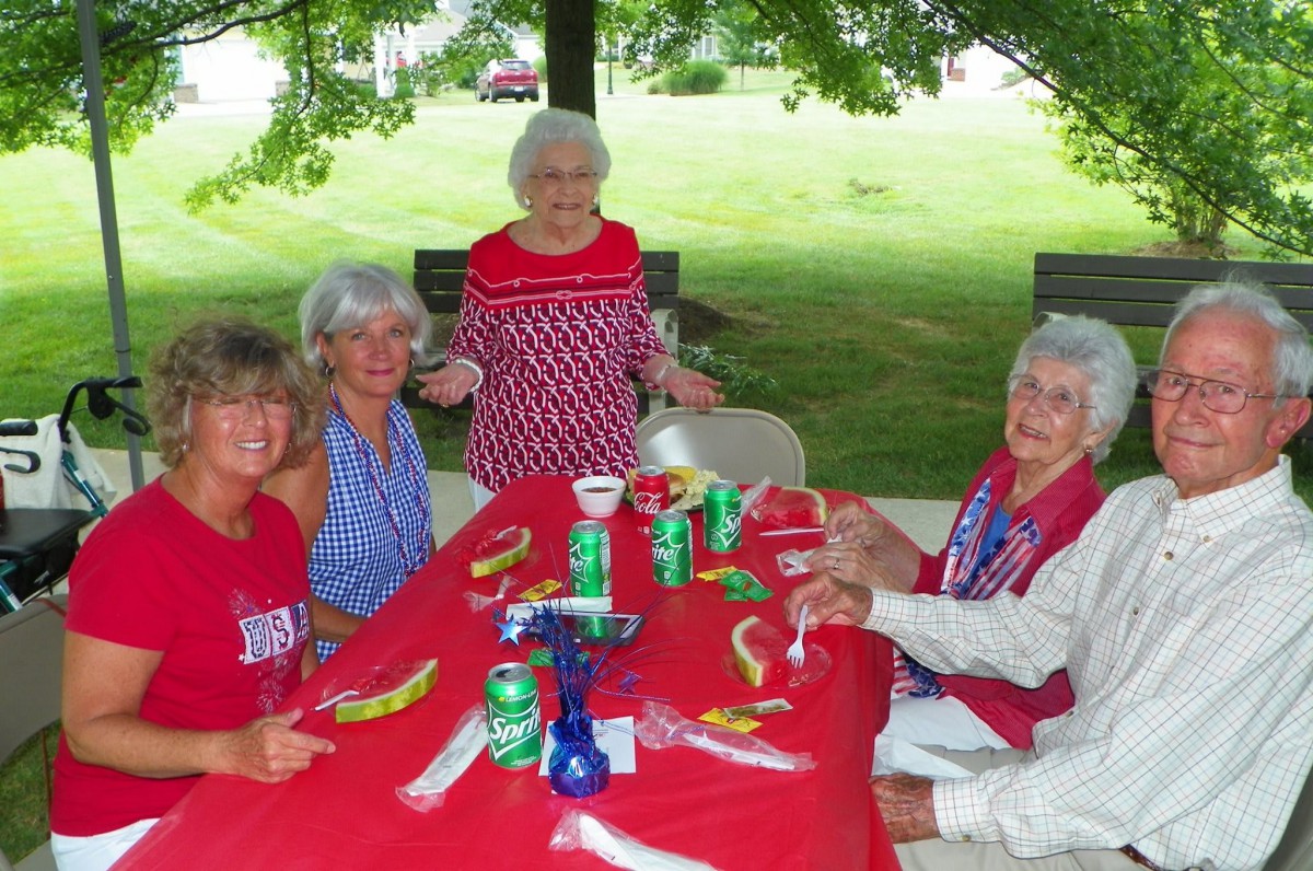 A group of people enjoying an outdoor picnic with food and drinks on a red tablecloth.