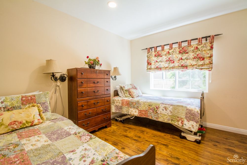 A cozy bedroom with twin beds featuring quilted bedspreads, a wooden dresser, and floral curtains over the window.