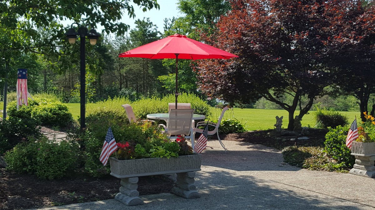 A peaceful garden scene with a red umbrella over patio seating, surrounded by greenery and small flags.