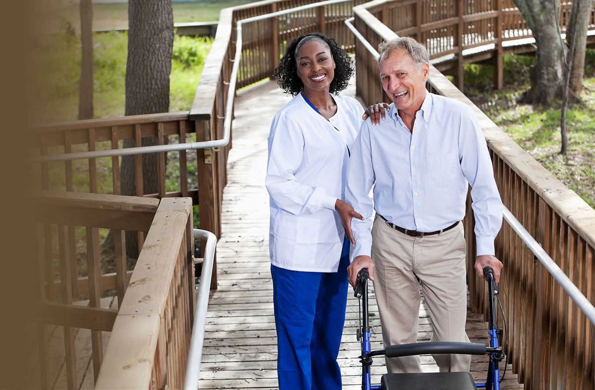 A nurse assisting an elderly man using a walker on a wooden path outdoors.
